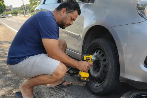 Motorista usando chave de impacto a bateria 21V para soltar parafuso de roda de carro na rua com macaco hidráulico
