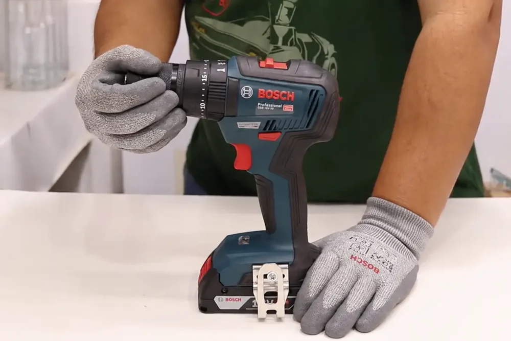 Parafusadeira Bosch GSB 18V-50 Professional segurada com luvas cinza, com bateria acoplada, sobre bancada branca